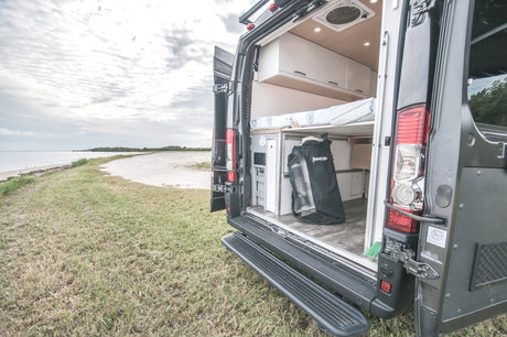 This RV rental camper van's open rear shows a comfy bed & gear storage, set on a grassy beach shore.