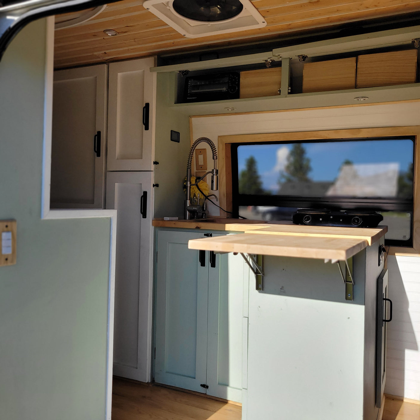 Inside an RV rental: a compact, functional kitchen with light wood cabinets, sink, window, and folding table in a custom van conversion.