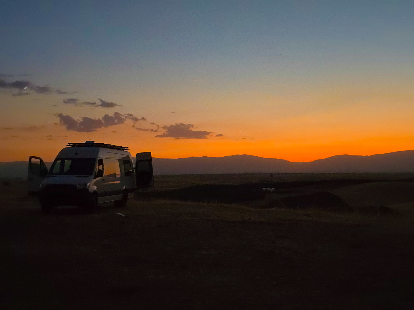 Serene sunset with an RV rental van in a field, mountains, and a crescent moon in the sky.
