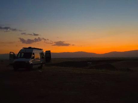 Serene sunset with an RV rental van in a field, mountains, and a crescent moon in the sky.