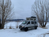 Experience winter adventures with an RV rental! A grey camper van with a pop-top tent is parked on snow beside a frozen lake & mountains.
