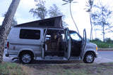 This Ford Econoline-style camper van, an ideal RV rental, has its doors open at a tropical park, revealing the comfy interior.