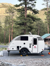 White Little Guy teardrop travel trailer, ideal for an RV rental, parked amidst tall pine trees at a mountain campsite.