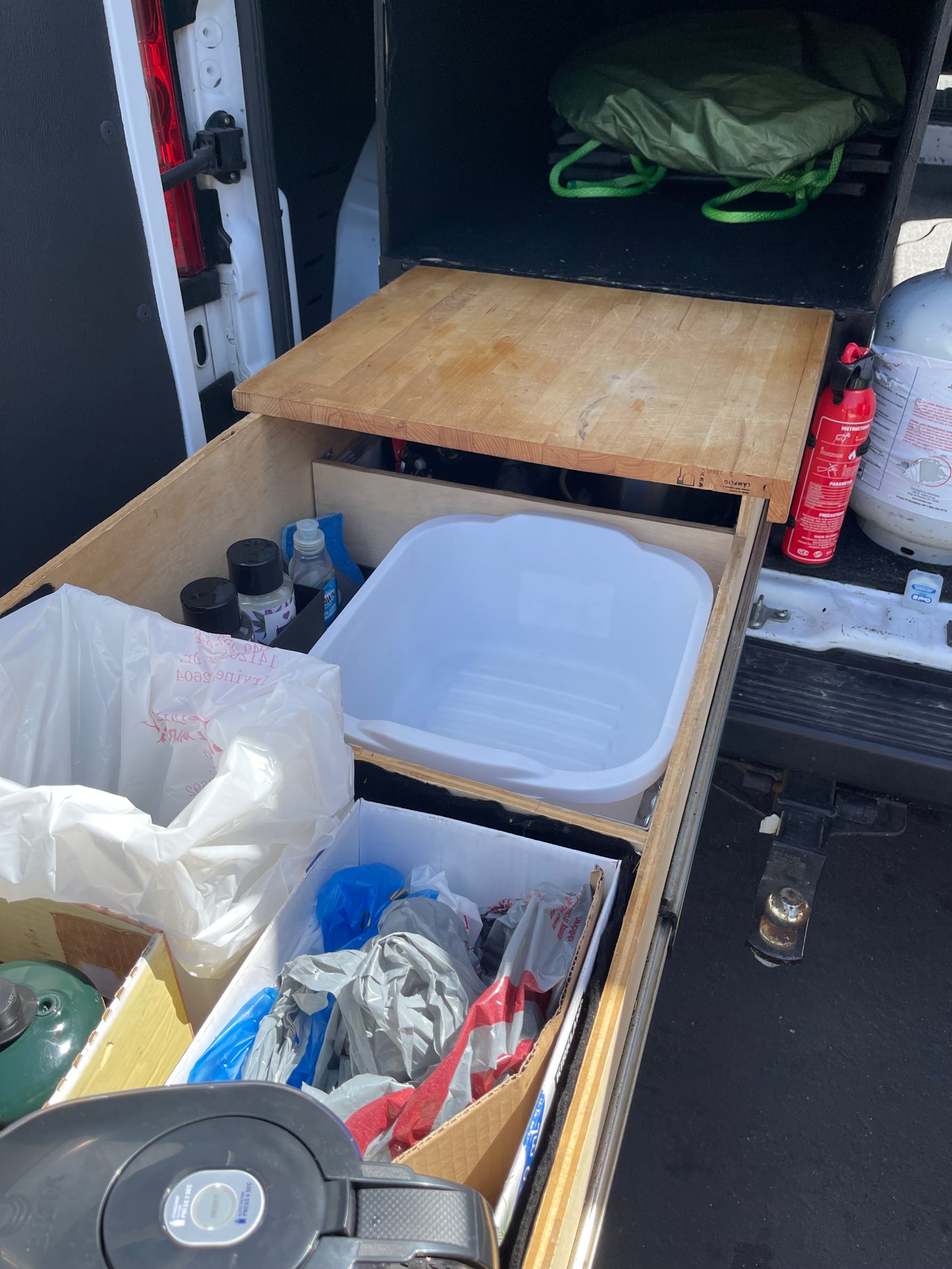 Organized rv rental pull-out kitchen/storage in a van: basin, cutting board, fire extinguisher, cleaning supplies, & camping gear.