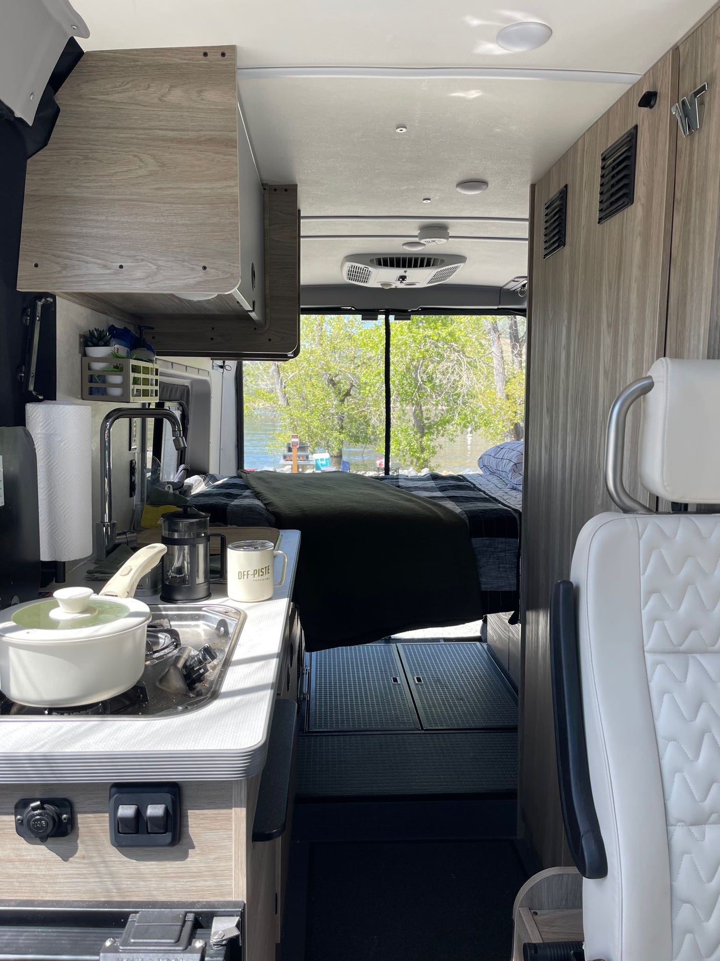 Cozy RV rental: compact kitchen with stove, sink, and coffee, plus a comfortable bed overlooking a serene river.