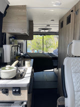Cozy RV rental: compact kitchen with stove, sink, and coffee, plus a comfortable bed overlooking a serene river.