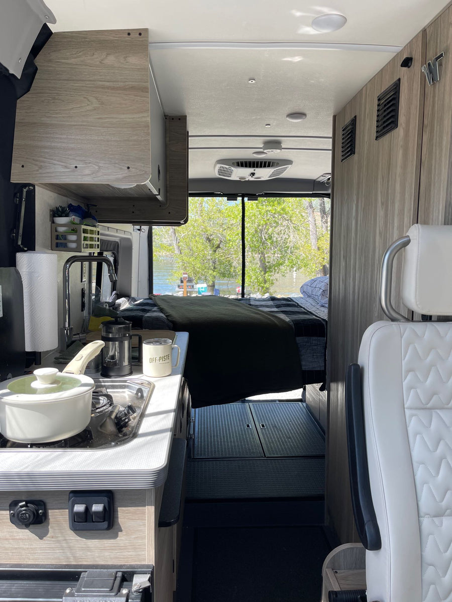 Cozy RV rental: compact kitchen with stove, sink, and coffee, plus a comfortable bed overlooking a serene river.