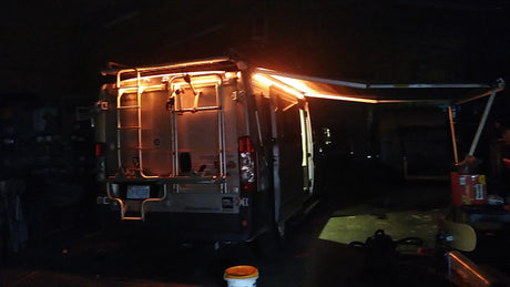 Night shot of an RV rental van with its rear lights and extended awning illuminated in a dark garage.
