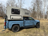 Light blue Toyota Tacoma with a Scout truck camper, perfect for an rv rental adventure. Parked outdoors amidst bare trees.
