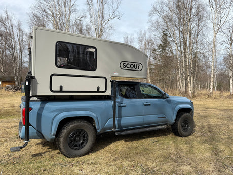 Light blue Toyota Tacoma with a Scout truck camper, perfect for an rv rental adventure. Parked outdoors amidst bare trees.