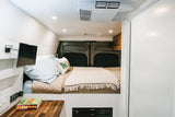 Clean RV rental interior: cozy bed, wall-mounted TV, countertop, built-in fridge, and overhead AC unit.