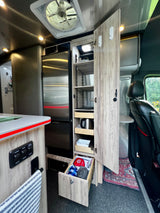 This modern RV rental interior features a stainless steel fridge, an open pantry with pull-out drawers, controls, and a driver's seat.