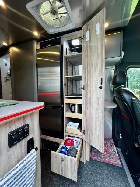 This modern RV rental interior features a stainless steel fridge, an open pantry with pull-out drawers, controls, and a driver's seat.