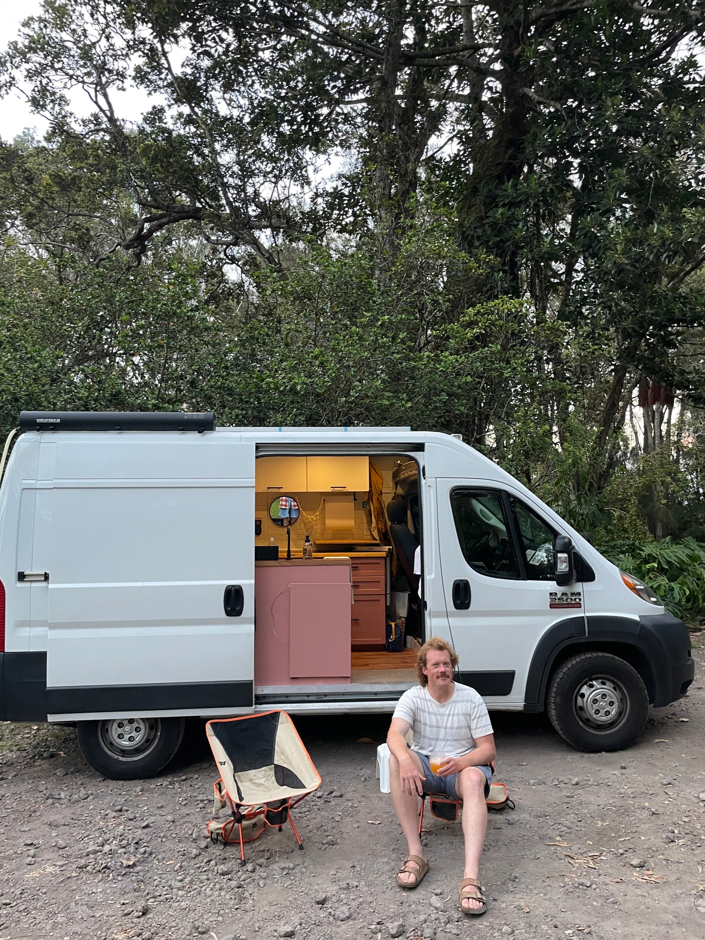 Man relaxing by an RV rental, a custom white camper van with a pink and white interior, parked among lush trees.