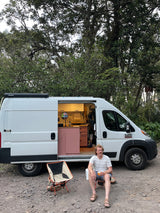Man relaxing by an RV rental, a custom white camper van with a pink and white interior, parked among lush trees.