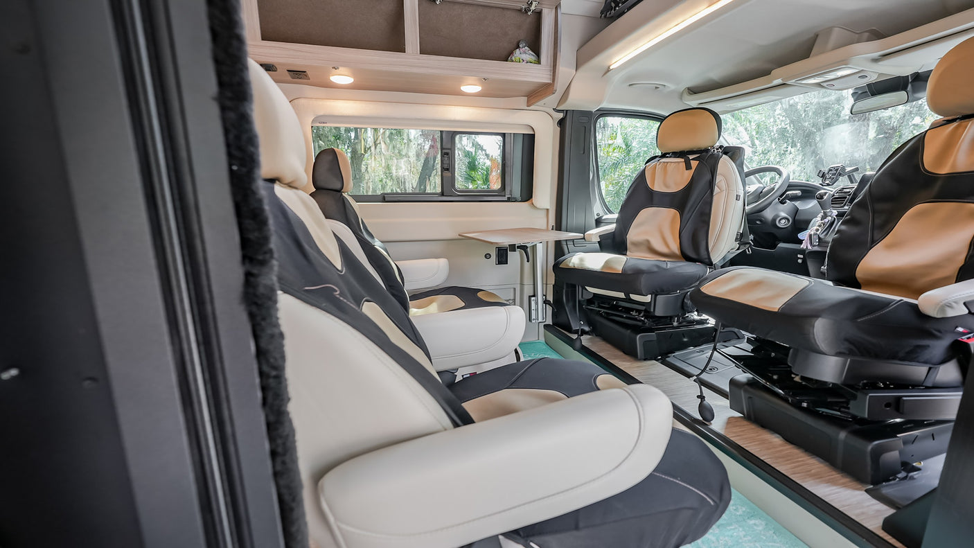 Interior of an rv rental with comfortable tan and black swivel seats, a small table, and overhead storage.