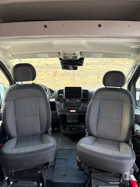 Inside an RV rental: spacious and clean front cabin with driver and passenger seats, modern dashboard, and open road view.