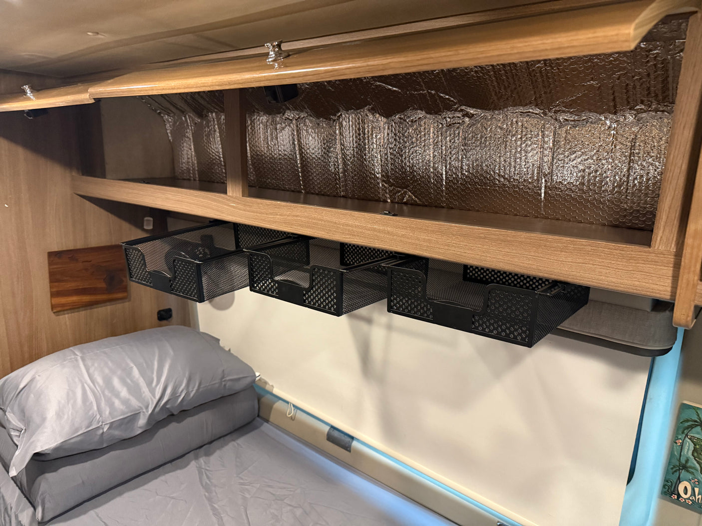 Overhead storage solutions in an RV rental: mesh baskets hang below a shelf, above a cozy bed, with reflective insulation visible.