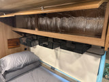 Overhead storage solutions in an RV rental: mesh baskets hang below a shelf, above a cozy bed, with reflective insulation visible.