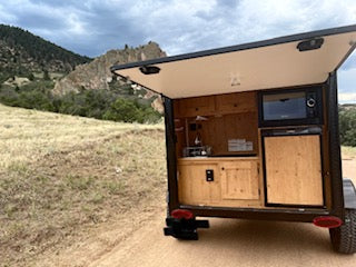RV rental: Compact camper trailer with rear hatch open, revealing a full kitchen including microwave and fridge, parked in mountains.