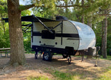 RV rental: A travel trailer with its awning extended is parked at a sunny, wooded campsite, a large tree on the left.
