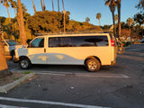 White RV rental van with a roof rack and bike, bathed in golden light, parked among tall palm trees. Perfect for trips!