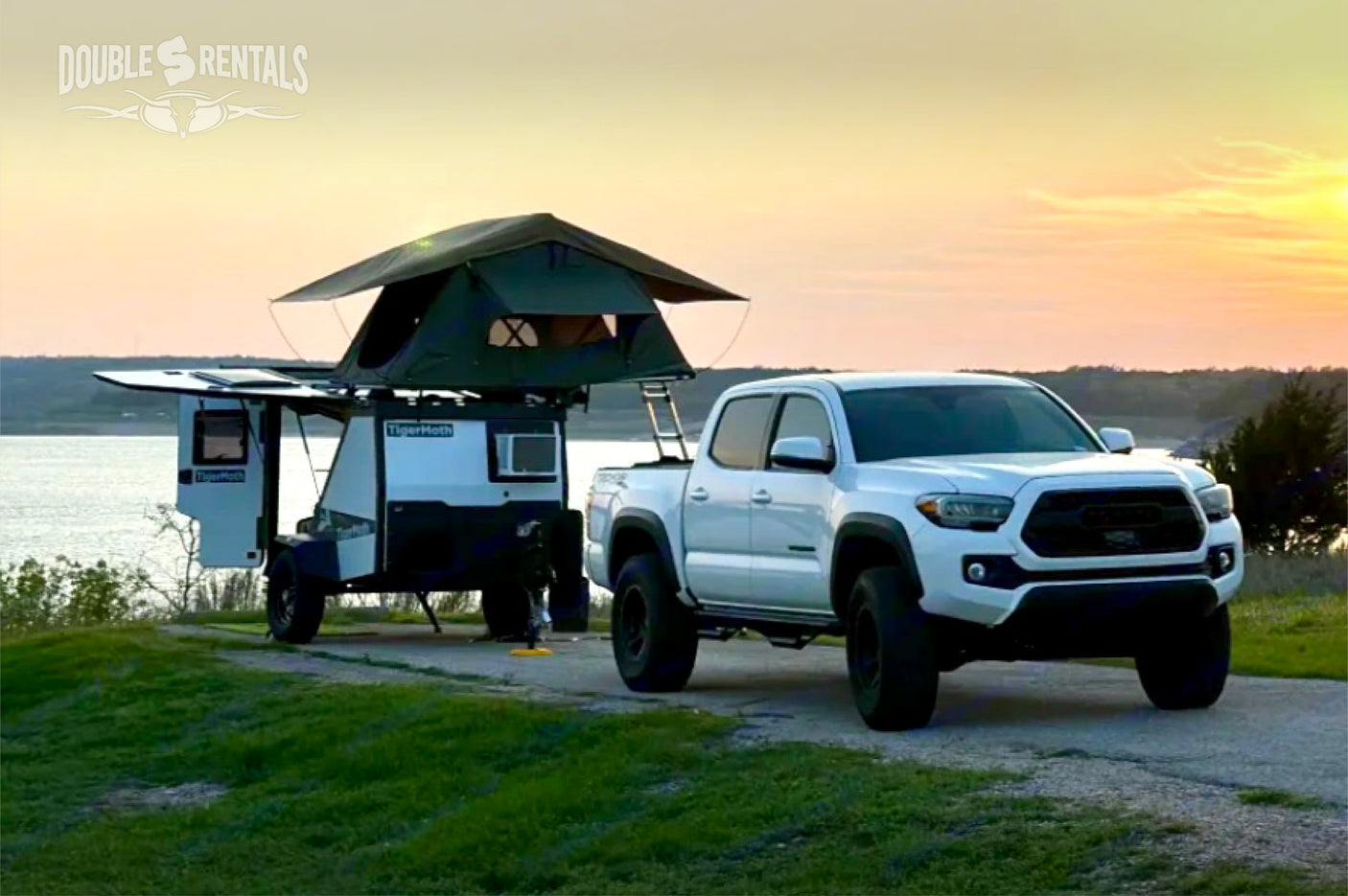 Experience adventure with this RV rental: White pickup, TigerMoth trailer & rooftop tent by a lake at sunset.