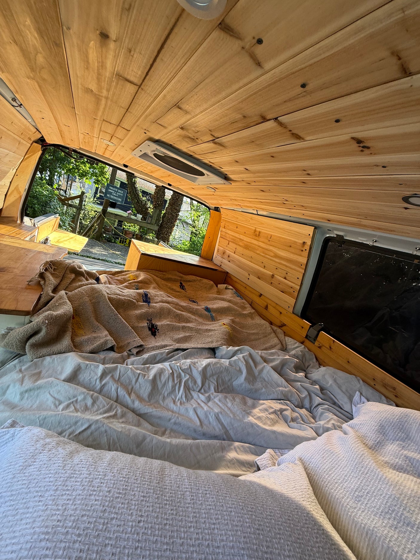 Explore comfort in this rv rental. Inside, a cozy bed and wood paneling. Outside, a lush view of trees & a 'No Trespassing' sign.