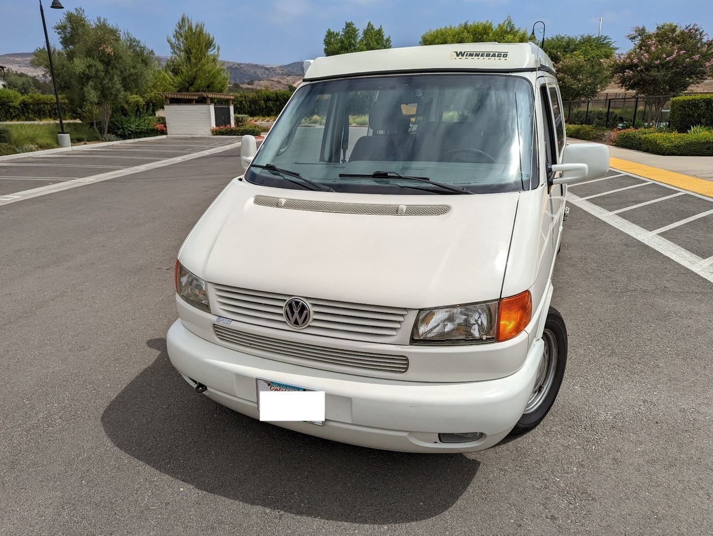 White Winnebago VW camper van, ideal for your next RV rental adventure. Parked on asphalt with trees & hills in the background. — photo 16