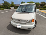 White Winnebago VW camper van, ideal for your next RV rental adventure. Parked on asphalt with trees & hills in the background.