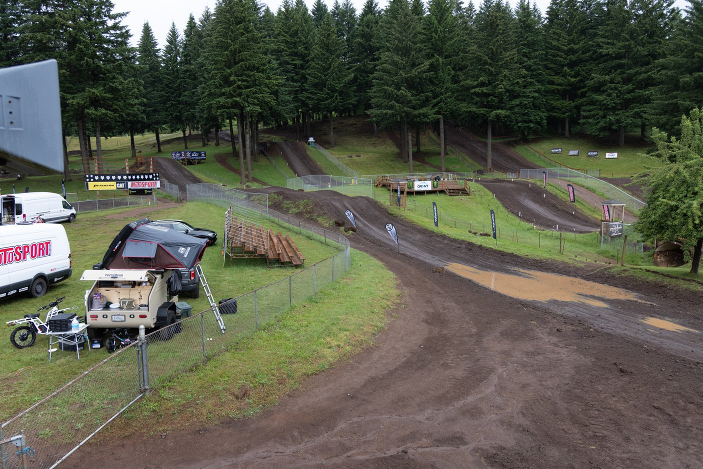 Muddy motocross track winds through dense trees. An RV rental-style trailer setup and vans are parked trackside for the event.