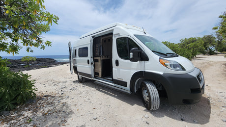 White RV rental camper van on a sandy, rocky beach. Ocean and volcanic shoreline in background. Perfect for nature adventures.