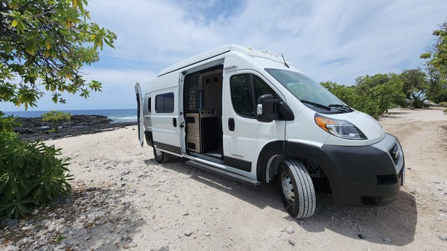 White RV rental camper van on a sandy, rocky beach. Ocean and volcanic shoreline in background. Perfect for nature adventures.