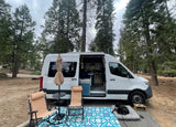 Serene forest scene with an RV rental camper van showing its open interior, outdoor seating, and a dog relaxing.