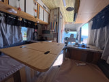 Bright rv rental interior featuring a spacious fold-out table, kitchen area with sink/stove, and beautiful views through the windows.