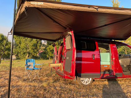 A red camper van RV rental with an open side, revealing a custom interior, parked under an awning with a chair outside.