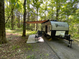 Viking Max travel trailer rv rental set up at a wooded campsite with awning, chairs, and rug, offering outdoor relaxation.