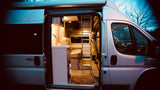 An RV rental from General RV Center, side door open to reveal a bright interior with kitchen, seating, & loft bed.