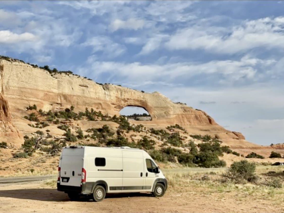 An RV rental camper van is parked on dirt near a natural sandstone arch in a vast desert landscape under a cloudy sky.|Explore nature with this Ram ProMaster RV rental. White van parked on a dirt road