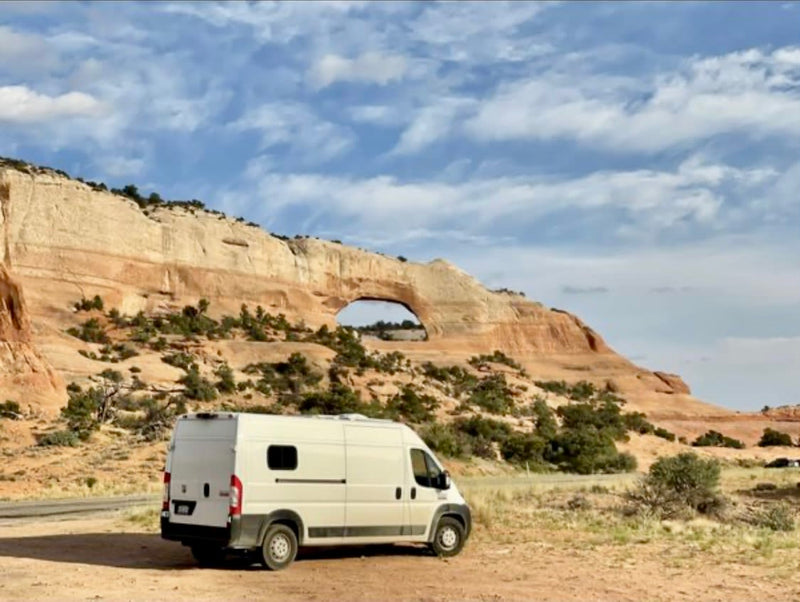 An RV rental camper van is parked on dirt near a natural sandstone arch in a vast desert landscape under a cloudy sky.|Explore nature with this Ram ProMaster RV rental. White van parked on a dirt road — photo 1