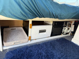 Under-bed storage in an RV rental: Dometic fridge, microwave, propane tank, and woven basket. Blue bedding and shag rug.