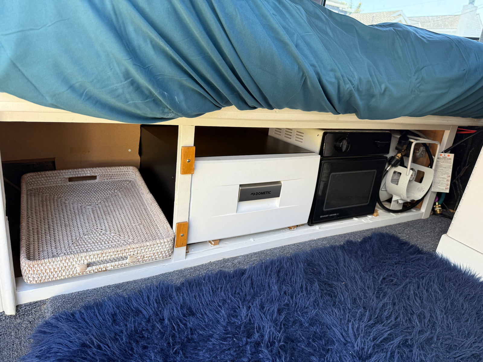 Under-bed storage in an RV rental: Dometic fridge, microwave, propane tank, and woven basket. Blue bedding and shag rug.