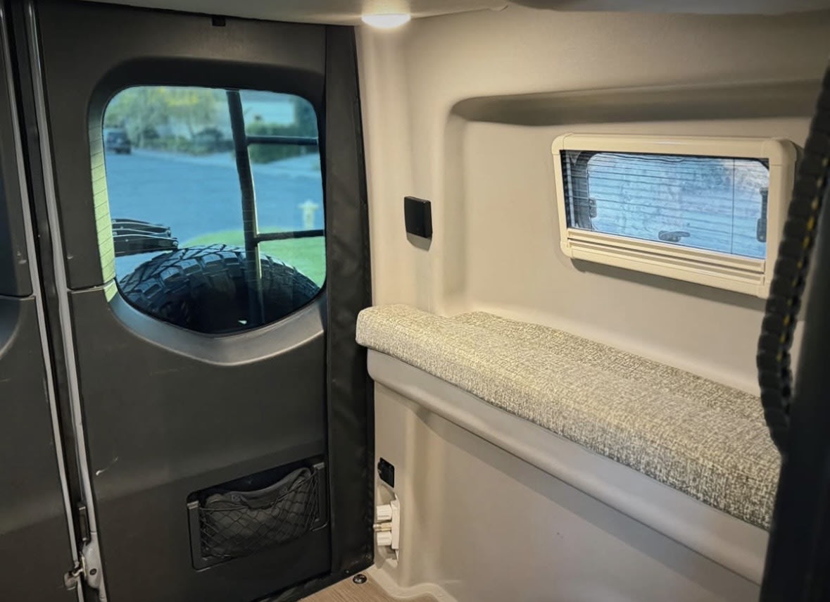 An RV rental's inviting interior: rear door window shows a mounted tire, a comfy bench, and a side window with blinds. — photo 16