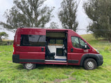 Red Ram ProMaster camper van with open side door showing custom interior for an RV rental adventure in a grassy field.