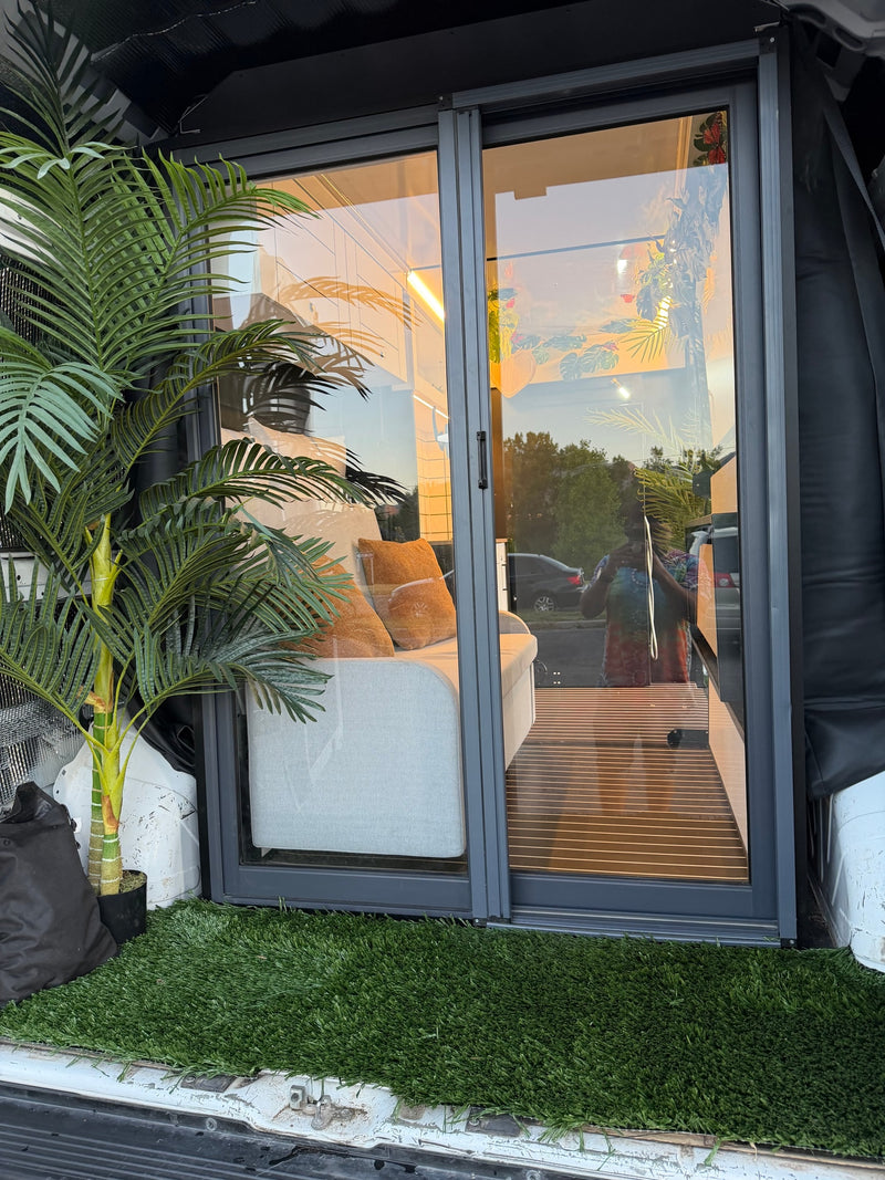 Compact rv rental living area seen through sliding glass doors, featuring a sofa, artificial palm plant, and outdoor grass mat. — photo 12