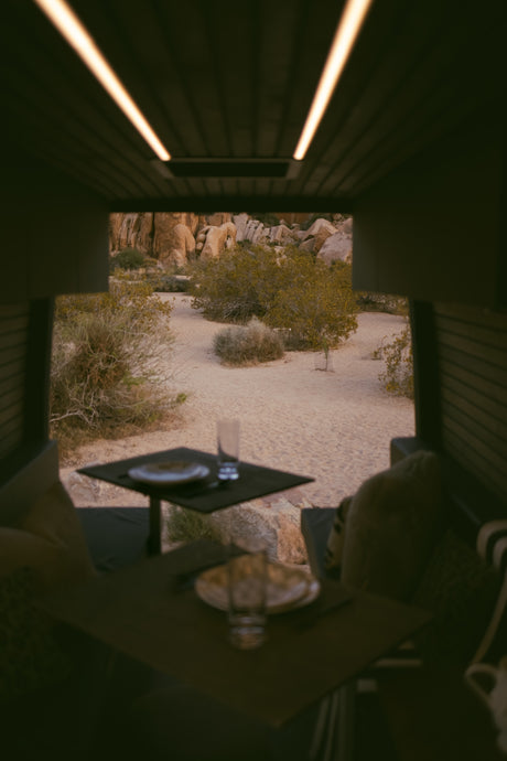 RV rental dining area, cozy interior with ceiling lights, framing a beautiful Joshua Tree desert landscape of rocks & bushes.