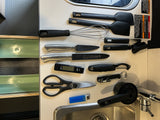 An RV rental kitchen counter organized with essential cooking tools: spatulas, whisk, knives, measuring cups, and a digital thermometer.