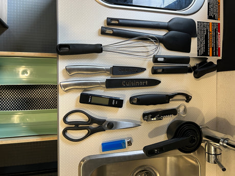 An RV rental kitchen counter organized with essential cooking tools: spatulas, whisk, knives, measuring cups, and a digital thermometer.