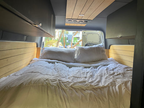 Comfortable RV rental interior with a cozy bed, white bedding, wooden accents, and open rear doors revealing a scenic view.