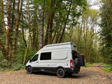 Light gray RV rental van with off-road tires, parked on a forest path amid moss-covered trees, ready for adventure.